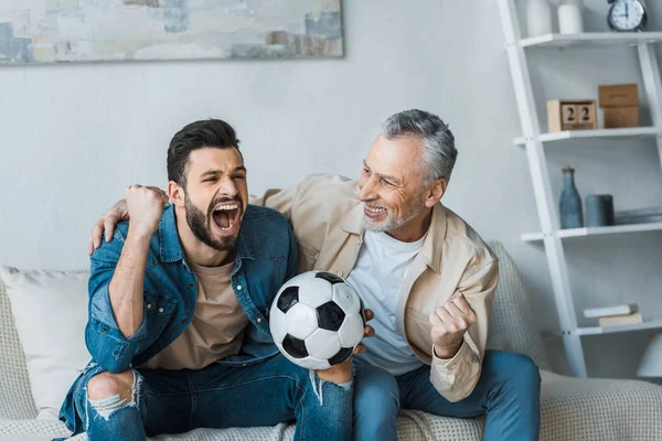 Älterer Mann schaut glücklichen Sohn beim Jubeln an, während er die Meisterschaft anschaut und Fußball hält — Stockfoto