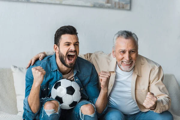 Glücklicher älterer Mann schaut Meisterschaft zu und jubelt mit hübschem Sohn, der Fußball hält — Stockfoto