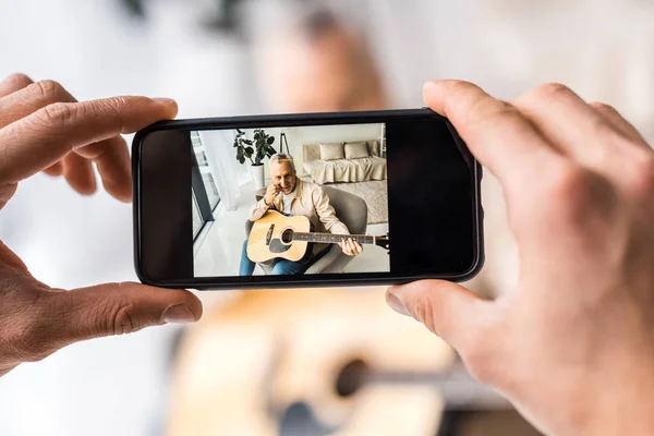 Vue recadrée de l'homme prenant une photo du père aîné tenant la guitare acoustique à la maison — Photo de stock