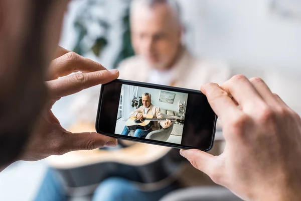 Abgeschnittene Ansicht eines Mannes, der ein Foto von einem Vater im Ruhestand macht, der zu Hause Akustikgitarre spielt — Stockfoto