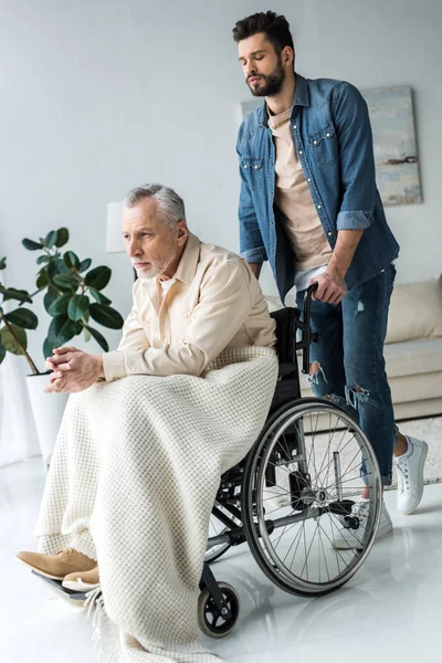 Guapo barbudo hijo sosteniendo silla de ruedas de discapacitados padre mayor en casa - foto de stock