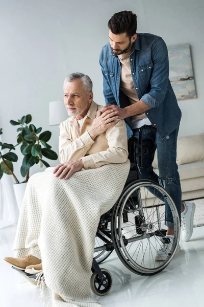 Beau fils barbu tenant la main avec un père aîné handicapé en fauteuil roulant à la maison — Photo de stock