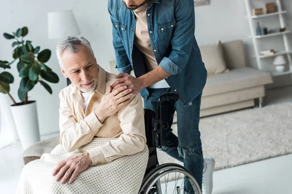 Vista ritagliata del figlio che si tiene per mano con il padre anziano disabile in sedia a rotelle a casa — Foto stock