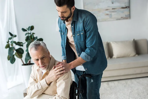 Caring handsome son holding hands with disabled senior father in wheelchair at home — Stock Photo
