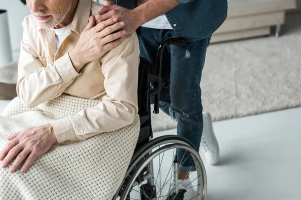 Обрезанный вид заботливого сына, держащегося за руки со старшим отцом-инвалидом в инвалидной коляске дома — стоковое фото