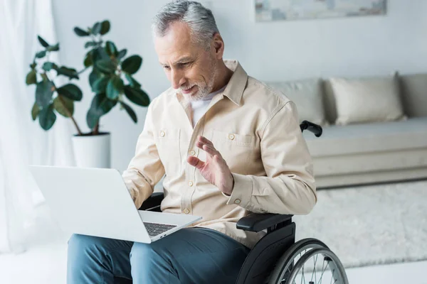 Веселый пенсионер-инвалид сидит в инвалидной коляске и машет рукой во время видеозвонка — стоковое фото