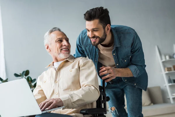 Веселый пенсионер-инвалид сидит в инвалидной коляске рядом с красивым сыном и смотрит на ноутбук — стоковое фото
