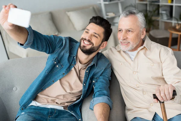 Figlio allegro prendendo selfie con padre allegro in pensione su smartphone — Foto stock