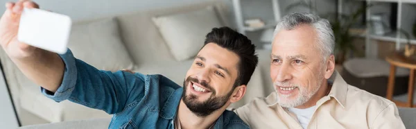 Plano panorámico de feliz barbudo hijo tomando selfie con jubilado alegre padre en casa - foto de stock