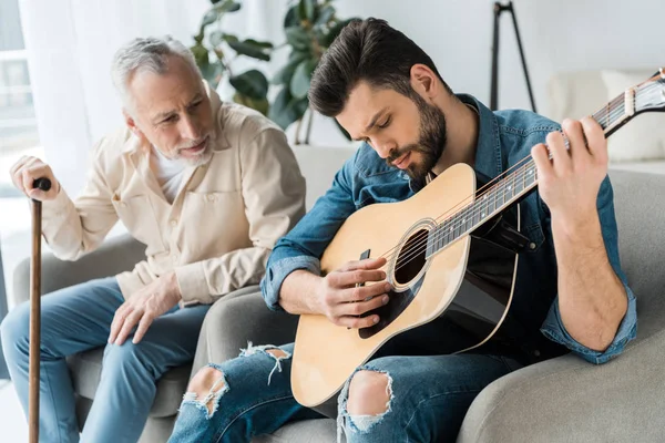 Guapo barbudo hombre tocando la guitarra acústica cerca feliz padre mayor en casa - foto de stock