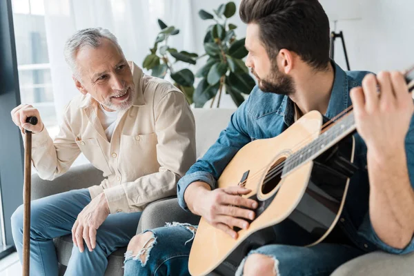 Бородатый сын играет на акустической гитаре рядом с счастливым старшим отцом дома — стоковое фото