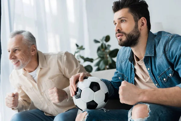 Felice pensionato guardando campionato e tifo con il figlio barbuto in possesso di calcio — Foto stock