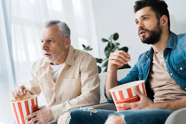 Conmocionado padre mayor sentado con su hijo y sosteniendo palomitas de maíz cubo mientras veía la televisión - foto de stock