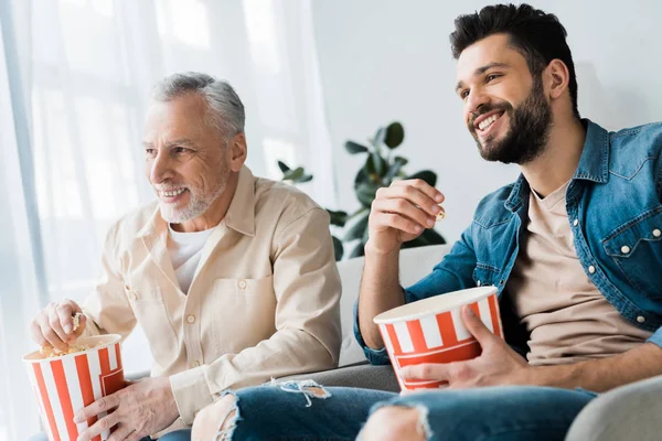 Feliz padre jubilado sentado con su hijo y sosteniendo palomitas de maíz cubo mientras mira la televisión - foto de stock