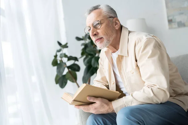 Hombre mayor en gafas sosteniendo libro mientras está sentado en casa - foto de stock