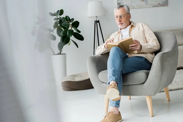 Pensativo hombre mayor en gafas sosteniendo libro mientras está sentado en sillón en casa - foto de stock