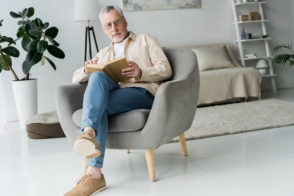 Pensive retired man in glasses holding book while sitting in armchair at home — Stock Photo