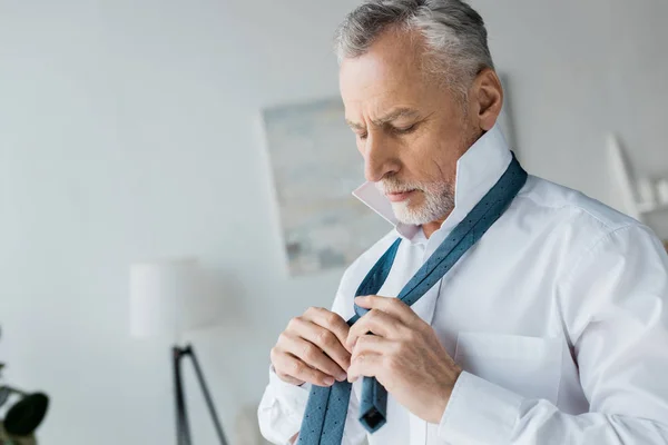 Elegant senior man tying tie while standing at home — Stock Photo