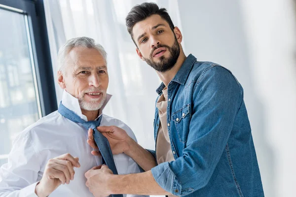 Handsome son helping cheerful retired father tying blue tie at home — Stock Photo