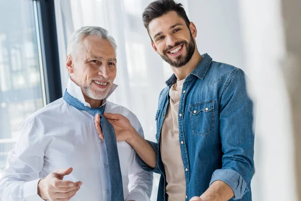 Happy son helping cheerful retired father tying blue tie at home — Stock Photo