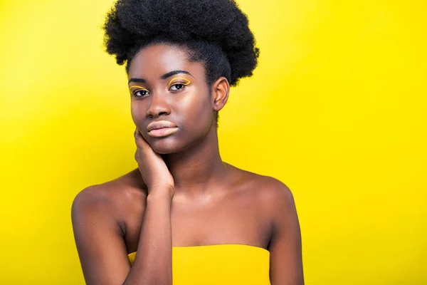 Atractiva mujer afroamericana con maquillaje de moda mirando a la cámara aislada en amarillo - foto de stock