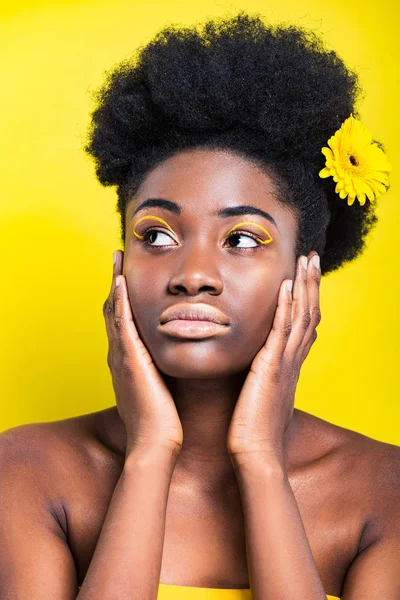 Мечтательная африканская американка с цветком в волосах, смотрящая вдаль, изолированная на желтом — стоковое фото
