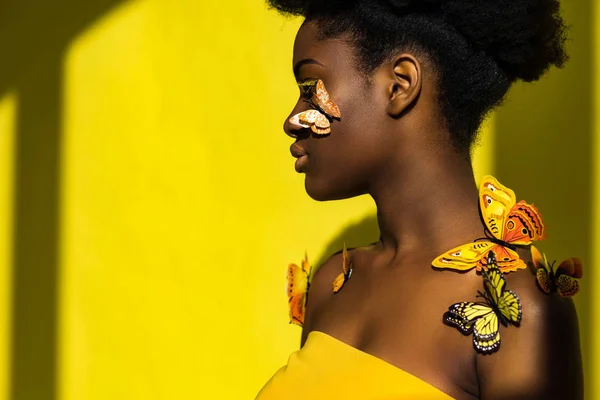 Vista lateral de mujer afroamericana con mariposas mirando hacia otro lado en amarillo - foto de stock