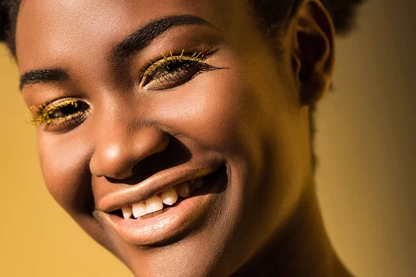 Улыбающаяся африканская американка с жёлтыми ресницами, смотрящая на камеру в коричневом. — стоковое фото