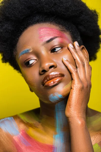 Mujer afroamericana pensativa con cuerpo arte tocando los labios y mirando hacia otro lado aislado en amarillo - foto de stock