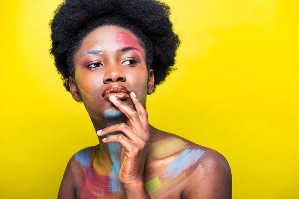 Mujer afroamericana pensativa con cuerpo arte tocando los labios y mirando hacia otro lado aislado en amarillo - foto de stock