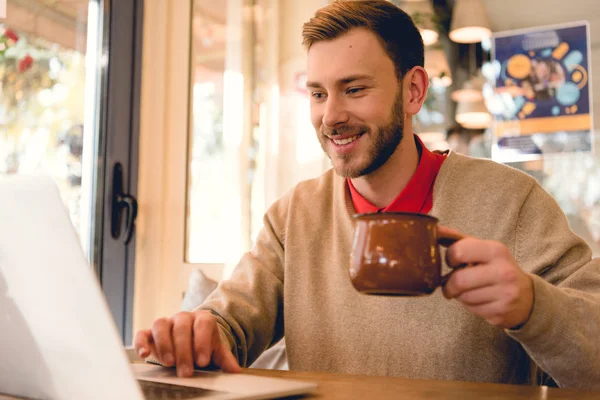 Blogueur barbu gai en utilisant un ordinateur portable et tenant une tasse de café dans le café — Photo de stock