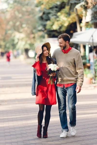 Uomo felice guardando la ragazza in cappello che tiene i fiori mentre cammina nel parco — Foto stock