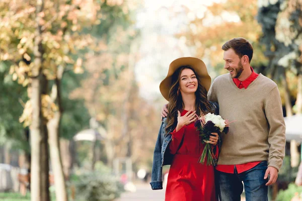 Happy man looking at girlfriend in hat smiling while holding flowers — Stock Photo