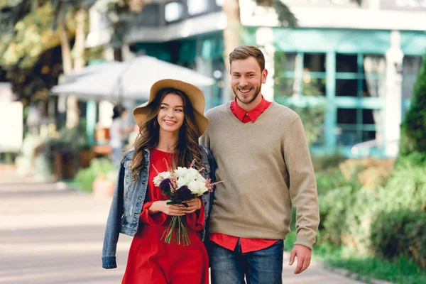 Happy man standing with attractive girlfriend in hat holding flowers — Stock Photo