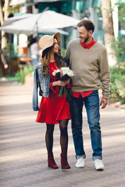 Heureux homme marche avec attrayant copine dans chapeau tenant des fleurs — Photo de stock