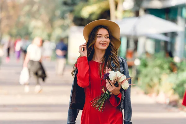 Glückliche Frau mit Hut lächelt, während sie Blumen im Park hält — Stockfoto
