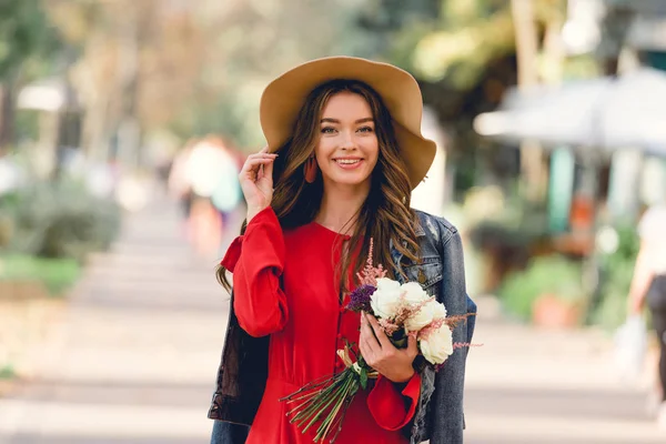 Mulher feliz no chapéu sorrindo enquanto segurando flores e olhando para a câmera — Fotografia de Stock