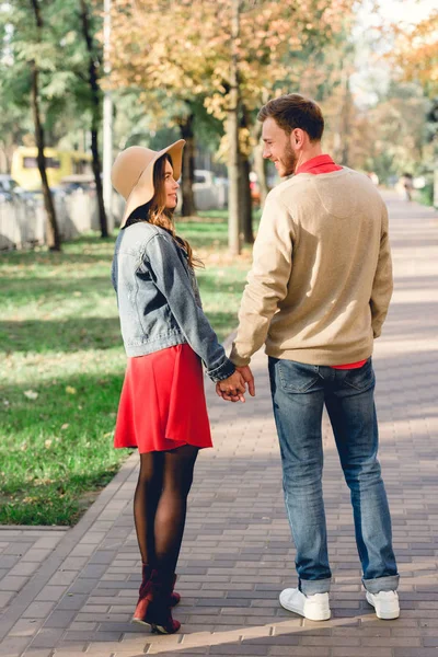 Handsome man looking at happy girlfriend in hat while holding hands in park — Stock Photo
