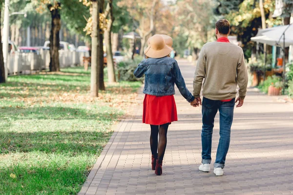 Vue arrière de l'homme et de la femme en chapeau tenant la main tout en marchant dans le parc — Photo de stock