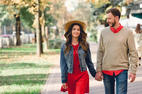 Fröhlicher Freund schaut glückliche Freundin mit Hut an, während er sich im Park an den Händen hält — Stockfoto