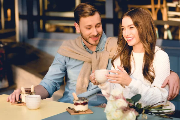 Cheerful man looking at cup in hands of happy girlfriend in cafe — Stock Photo