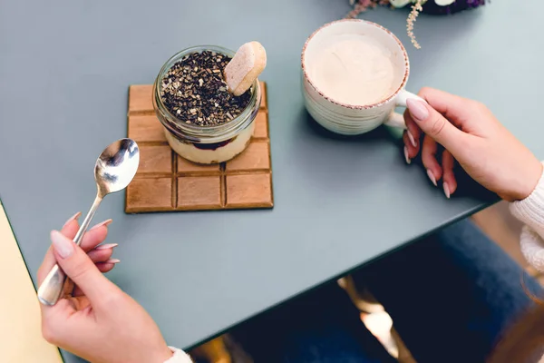 Vista ritagliata di giovane donna che tiene cucchiaio vicino a dessert saporito e tazza di caffè — Foto stock