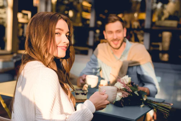 Enfoque selectivo de la chica feliz sosteniendo la taza con café mientras está sentado cerca del hombre en la cafetería - foto de stock