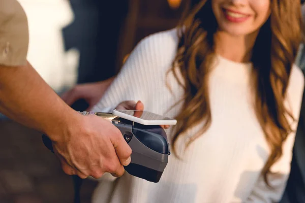 Vista ritagliata di donna allegra che paga con smartphone in caffè — Foto stock