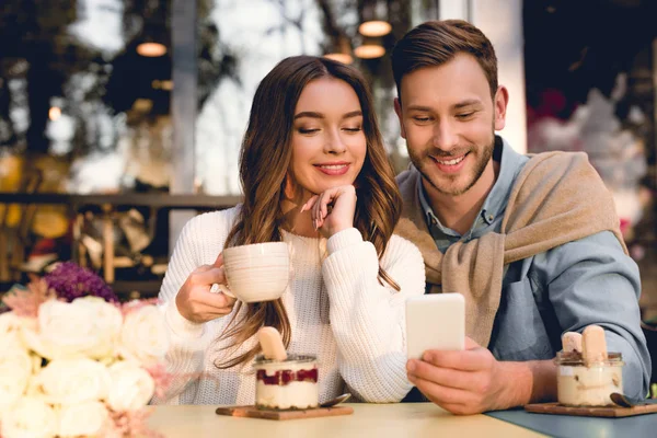 Homem alegre olhando para smartphone perto atraente menina segurando xícara de café — Fotografia de Stock