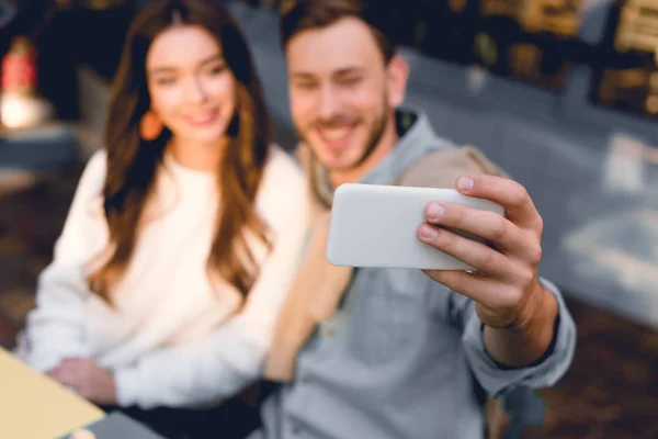 Selective focus of smartphone in hand of cheerful man taking selfie with young woman — Stock Photo