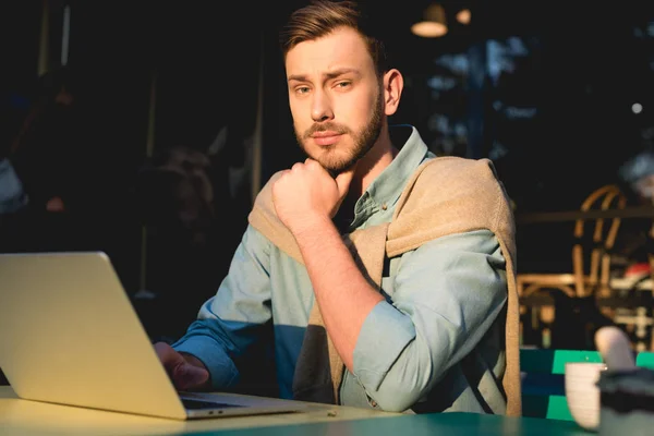 Pensive bearded freelancer sitting near laptop and looking at camera — Stock Photo