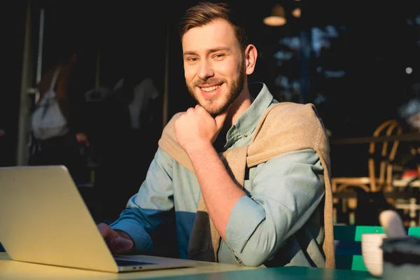 Fröhlicher bärtiger Freiberufler, der neben Laptop sitzt und in die Kamera blickt — Stockfoto