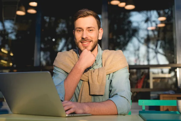 Cheerful and handsome freelancer sitting near laptop and looking at camera — Stock Photo