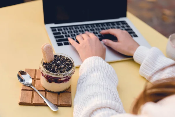 Cropped view of girl typing on laptop near sweet dessert and spoon — Stock Photo